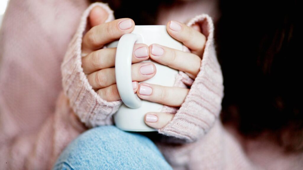 Close-up of hands holding a white mug, wrapped in a soft pink sweater—capturing a quiet moment of strength, stillness, and chronic illness resilience.