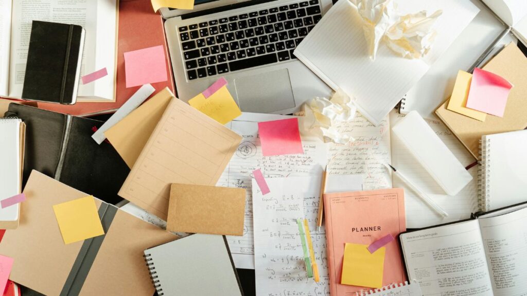 Overhead shot of a cluttered desk covered in planners, sticky notes, a laptop, and crumpled paper—capturing the mental chaos of chronic illness management.
